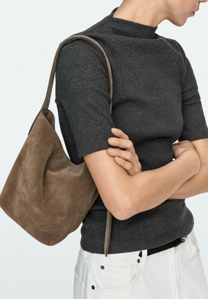 Persona che indossa una maglietta a maniche corte grigio scuro e pantaloni bianchi, con una borsa a tracolla in camoscio marrone, a braccia incrociate.