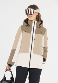 Giacca da sci beige e bianca, con cappuccio, chiusura con zip e blocchi di colore a contrasto. Una persona indossa occhiali da sci specchiati e tiene un casco.