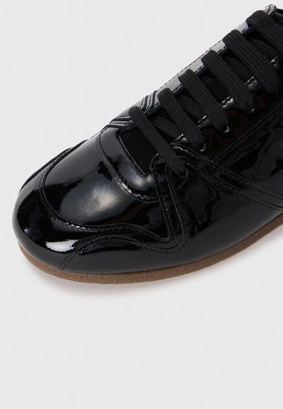Glossy black leather sneaker with black laces and light brown rubber sole, shown in close-up of the front and side.