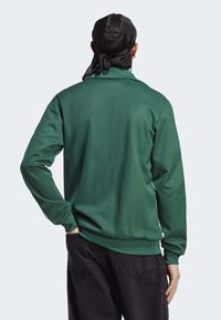 Grön sweatshirt med långa ärmar, slät textur, ribbade ärmslut och nederkant, bärs över svarta byxor, sedd bakifrån.