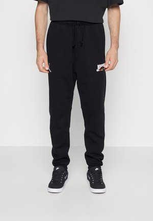 Mand iført sorte Jordan joggingbukser og sorte Nike sneakers, stående mod en ensfarvet lys baggrund.