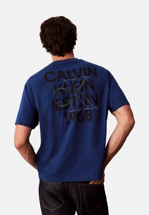 Hombre que lleva una camiseta azul de Calvin Klein con texto de logo en negro y blanco en la espalda, de pie con una mano en el bolsillo.