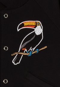 Tessuto nero con un tucano ricamato in dettaglio posato su un ramo, dotato di un becco colorato, zampe blu e accenti bianchi.