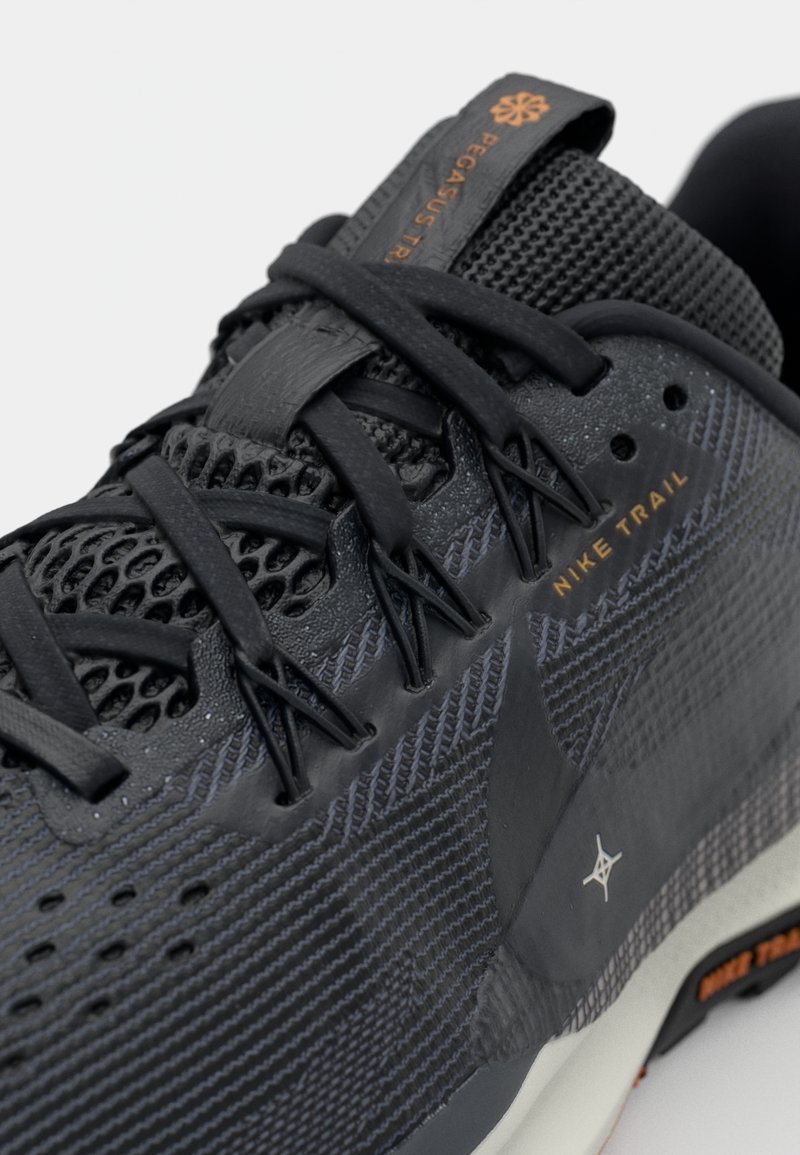 Zapatilla de trail running negra de Nike con parte superior de malla texturizada, puntera reforzada y detalles naranjas en la lengua y el lado.