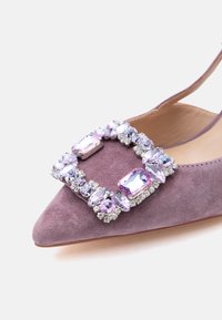 Μοκασίνι από σουέντ σε λιλά απόχρωση με μυτερή μύτη και τετράγωνη αγκράφα διακοσμημένη με ανοιχτόμωβ και διαφανείς πολύτιμους λίθους.