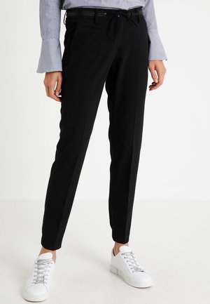 Czarne, dopasowane spodnie z wąskim krojem i ściągaczem w talii, noszone z białymi tenisówkami i koszulą w paski z rozszerzanymi mankietami na modelce stojącej.