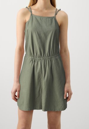 Femme portant une robe en lin sans manches de couleur olive avec de fines bretelles et une taille élastique, debout devant un fond clair uni.