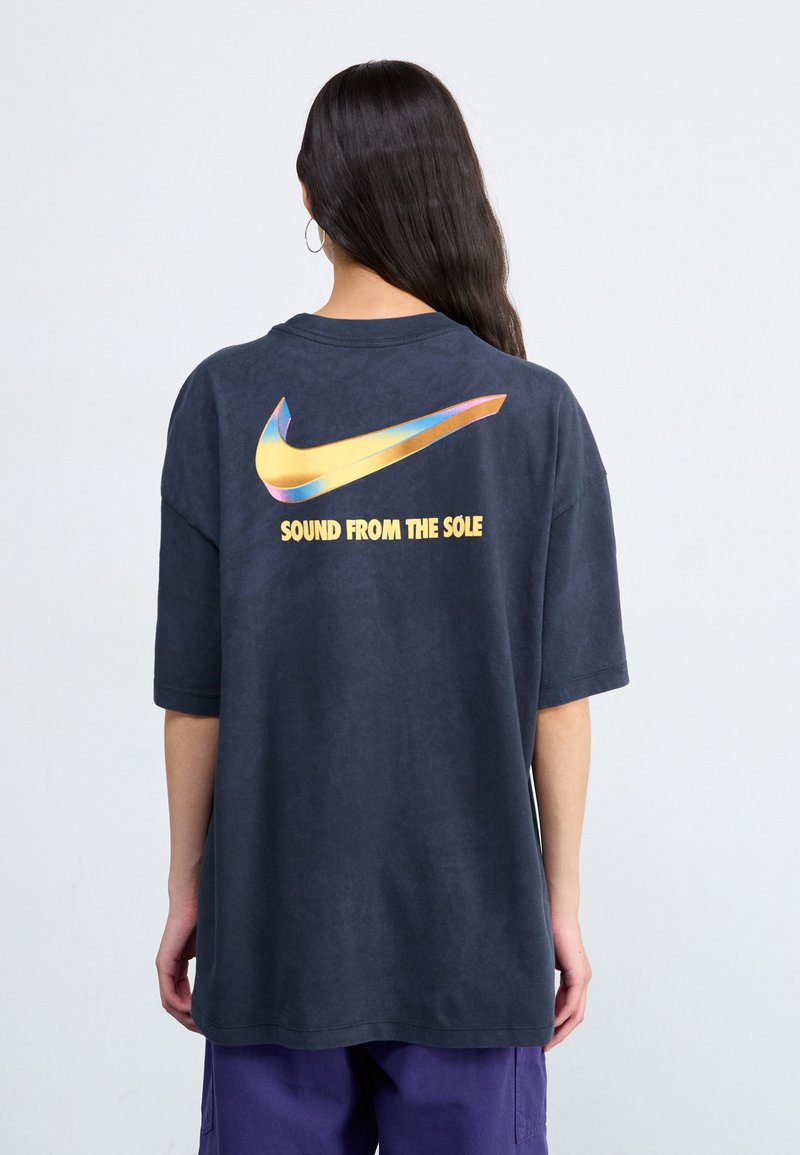 Temno siva oversized Nike majica z velikim iridiscentnim logotipom in napisom "ZVOK IZ PODPLATA" na hrbtni strani. Gladka tekstura.