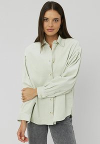 Chemise en velours côtelé vert clair avec un boutonnage à l'avant, manches longues et col classique. Présente des rayures verticales texturées et une coupe décontractée.