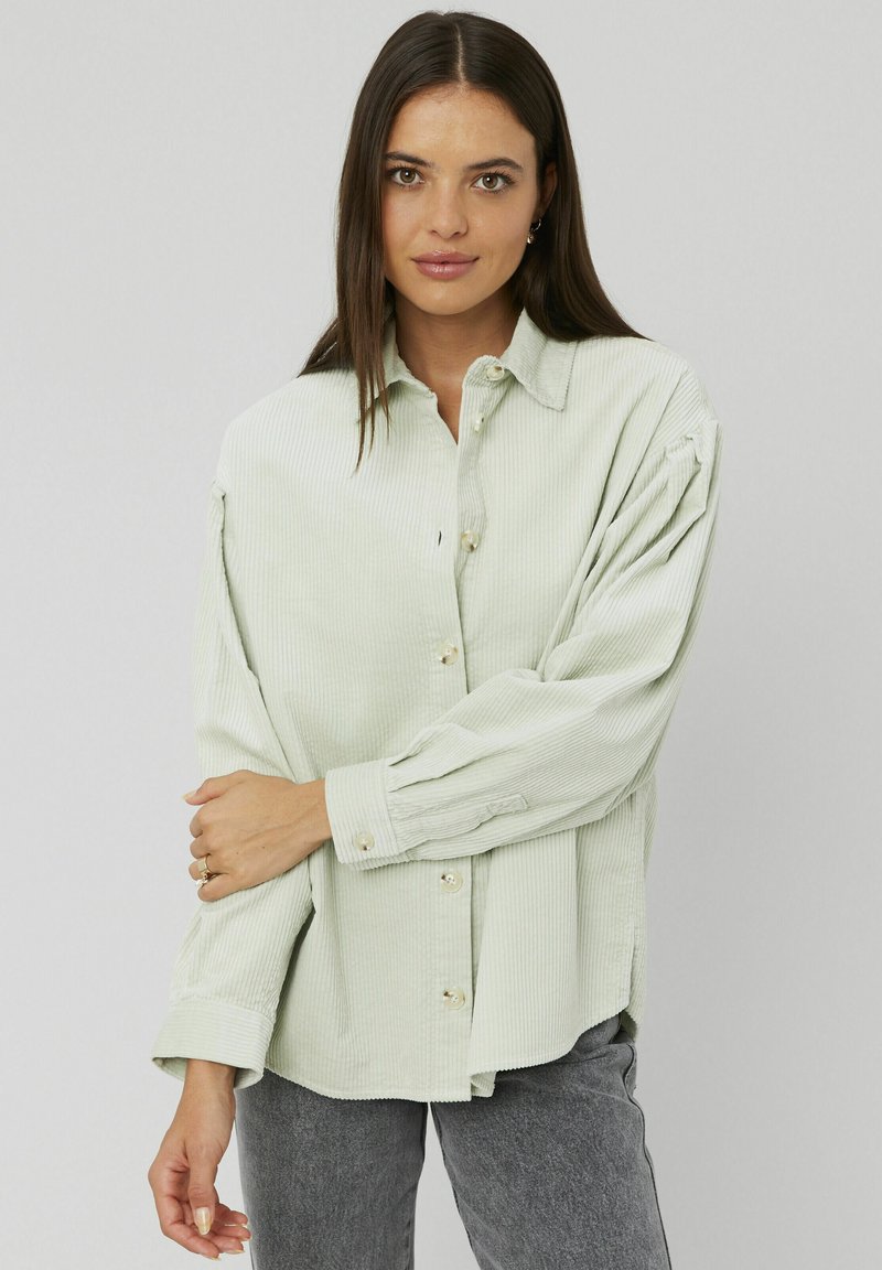 Chemise en velours côtelé vert clair avec un boutonnage à l'avant, manches longues et col classique. Présente des rayures verticales texturées et une coupe décontractée.