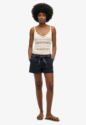 Canottiera bianca all'uncinetto con scollo a V, abbinata a pantaloni corti blu navy con una cintura a motivi. Il modello indossa sandali marroni. Sfondo semplice.