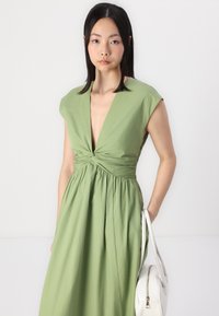 Abito verde con scollatura a V profonda, maniche a cappuccio e dettagli drappeggiati sul corpetto. Tessuto leggero che cade in modo fluido su una gonna plissettata. Borsa bianca.