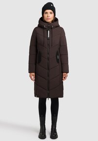 Vrouw die een lange zwarte gewatteerde jas met capuchon, zwarte beanie, zwarte Leggings en zwarte enkellaarsjes draagt, staande tegen een lichtgrijze achtergrond.