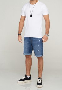 Hombre que lleva una camiseta blanca de manga corta, pantalones cortos de mezclilla desgastados y arremangados, zapatillas negras con suelas y cordones blancos, reloj, y collar con colgante negro.
