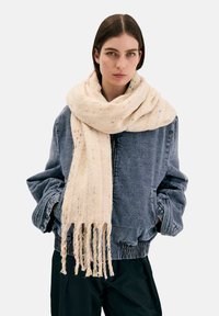 Giovane donna che indossa una giacca di jeans oversize e una grande sciarpa cremisi con macchie multicolori e margini sfrangiati, in piedi con le mani nelle tasche.