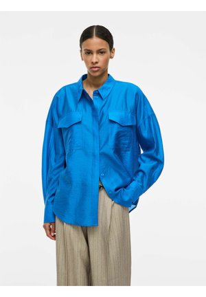 Kvinne med håret trukket tilbake, iført en skinnende blå, oversized skjorte med knapper og beige vidbukser med tynne striper, mot hvit bakgrunn.