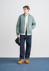 Πράσινο μπουφάν bomber με καπιτονέ υφή και μανσέτες με ριμπ, λευκό πουκάμισο από κάτω, σκούρο μπλε παντελόνι, κίτρινα βελούδινα μποτάκια και μαύρο καπέλο στο χέρι.