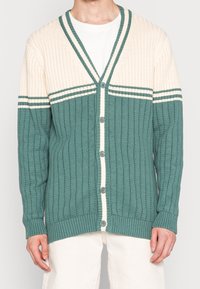 Cardigan acanalado en verde y crema con escote en V, cierre de botones y acentos de rayas horizontales. Patrón de punto texturizado en toda la prenda.