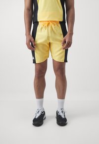 Żółte szorty sportowe z czarnymi panelami bocznymi, elastycznym pasem i sznurkiem do regulacji. Połączone z czarnymi butami i białymi skarpetkami sięgającymi do kostek.