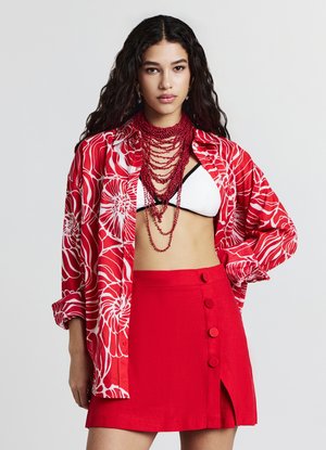 Chemise à boutons avec imprimé floral rouge, haut de bikini blanc, jupe rouge avec fente sur le côté et boutons, collier rouge superposé, cheveux texturés.