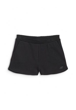 Dziewczyna stoi z rękami w kieszeniach, ubrana w jasnozielony luźny t-shirt, czarne szorty, białe skarpetki i fioletowe adidasy na szarym tle.