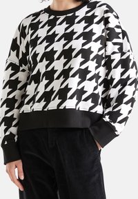 Person som bär en överdimensionerad sweatshirt i svart och vitt hounds-tooth mönster med svarta muddar och fåll, tillsammans med svarta kordflöjlsbyxor.