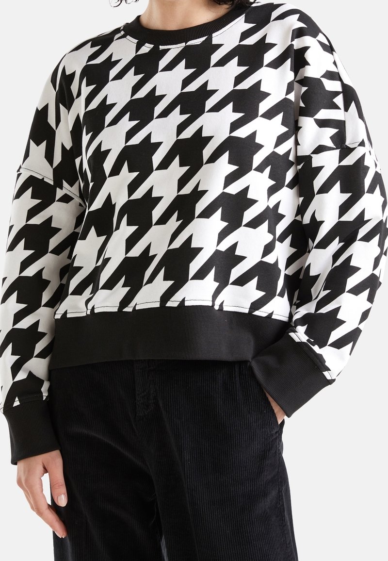 Person som bär en överdimensionerad sweatshirt i svart och vitt hounds-tooth mönster med svarta muddar och fåll, tillsammans med svarta kordflöjlsbyxor.