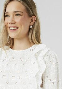 Blouse blanche brodée avec des accents froncés, présentant un motif floral et des manches bouffantes. Le modèle porte de grandes boucles d'oreilles métalliques.