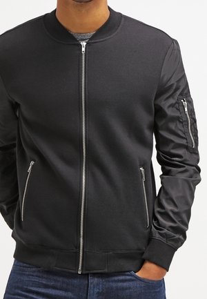 Homme portant une veste bomber noire avec fermeture éclair à l'avant, poches latérales zippées et poche zippée sur la manche gauche, assortie à un jean bleu.