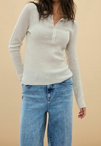 Lichtgrijs geribd langemouwen shirt met een henley halslijn en vijf knoopsluitingen. Gecombineerd met blauwe wijde jeans.