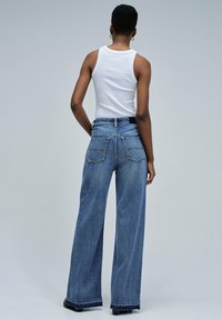 Person standing facing away wearing a white sleeveless top and wide-leg blue denim jeans with frayed hems.