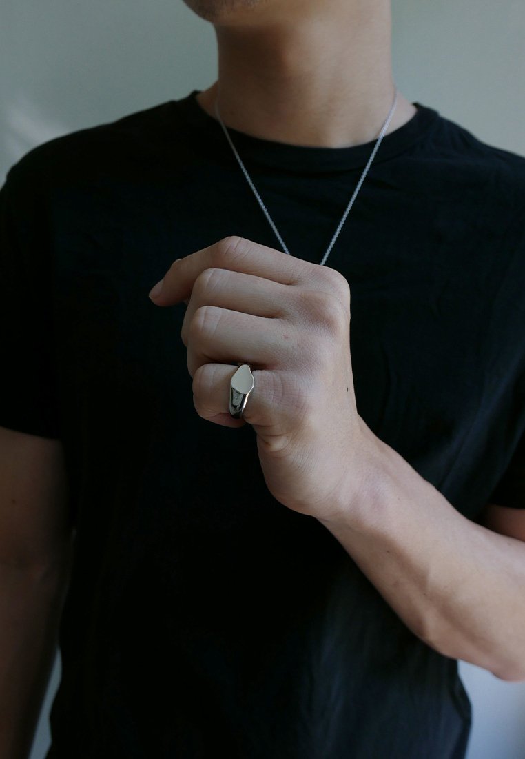 Silver ring with a smooth, elongated shape worn on a hand. The wearer is in a black shirt, with a simple silver necklace visible.