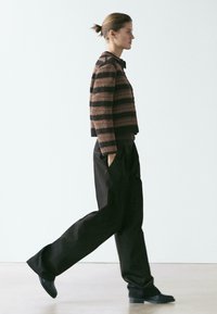 Frau geht seitlich, trägt einen braun-schwarz gestreiften Pullover, lockere schwarze Hose und schwarze Schuhe vor einem einfachen hellen Hintergrund.