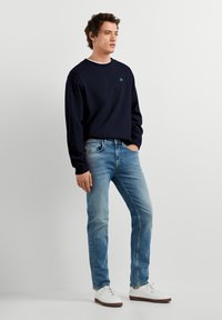 Jonge man staat met één hand in zijn zak, draagt een zwart shirt met lange mouwen, blauwe spijkerbroek en witte sneakers tegen een effen achtergrond.