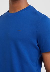 Detailní záběr osoby, která má na sobě jednoduché jasně modré tričko s malým logem "MK" na hrudi.