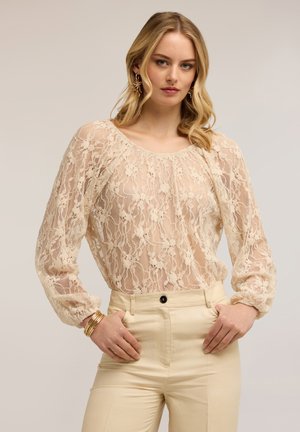 Lyshåret kvinde iført cremefarvet blonder-langærmet bluse og højtaljede beige bukser, poserende med hænderne på hofterne mod en ensfarvet baggrund.