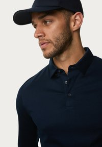 Marineblaues Langarm-Polohemd mit Knopfleiste und strukturiertem Kragen, getragen mit einer passenden marineblauen Kappe, die eine glatte Stoffstruktur aufweist.