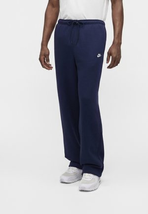 Homme portant un pantalon de jogging Nike bleu marine avec une taille à cordon et des baskets blanches, debout devant un fond clair uni.