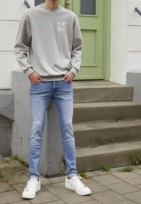 Grå sweatshirt med tryckt text, blå jeans och vita sneakers. I bakgrunden syns stensteg och en grön dörr.