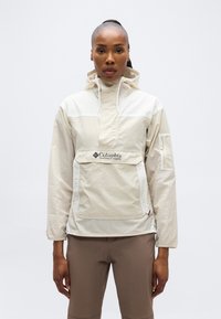 Γυναίκα που φοράει μπεζ αντιανεμικό μπουφάν Columbia Sportswear με μπροστινή τσέπη και κουκούλα, συνδυασμένο με καφέ παντελόνι, στέκεται μπροστά σε λευκό φόντο.