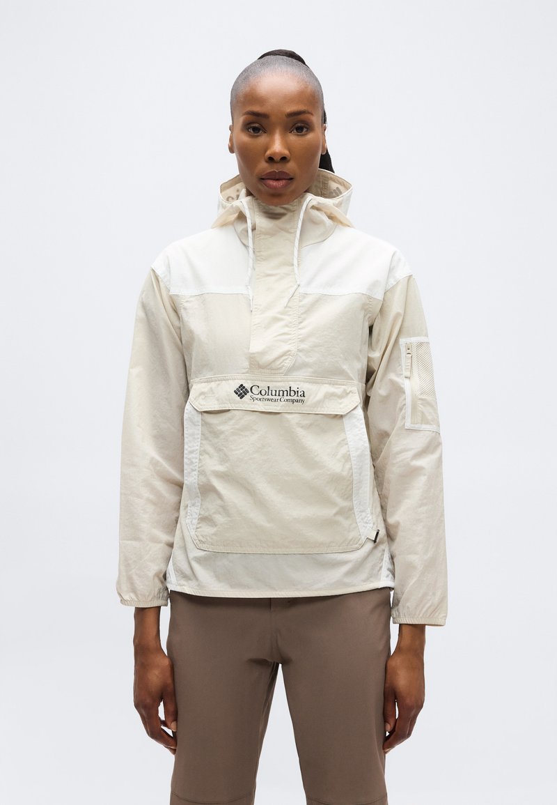 Γυναίκα που φοράει μπεζ αντιανεμικό μπουφάν Columbia Sportswear με μπροστινή τσέπη και κουκούλα, συνδυασμένο με καφέ παντελόνι, στέκεται μπροστά σε λευκό φόντο.