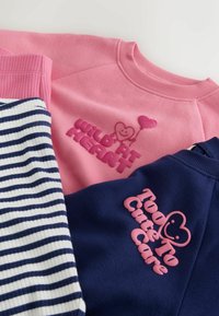 Vaaleanpunainen collegepaidassa teksti "Wild at Heart" ja sydän-ilmapallo, laivastonsininen collegepaidassa teksti "Too Cute To Care" ja hymyilevä sydän sekä raidalliset housut.