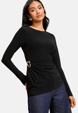 Top noir à manches longues avec un détail froncé sur le côté, accentué par une boucle circulaire en or. Tissu lisse avec une coupe ajustée. Associé à un jean foncé.