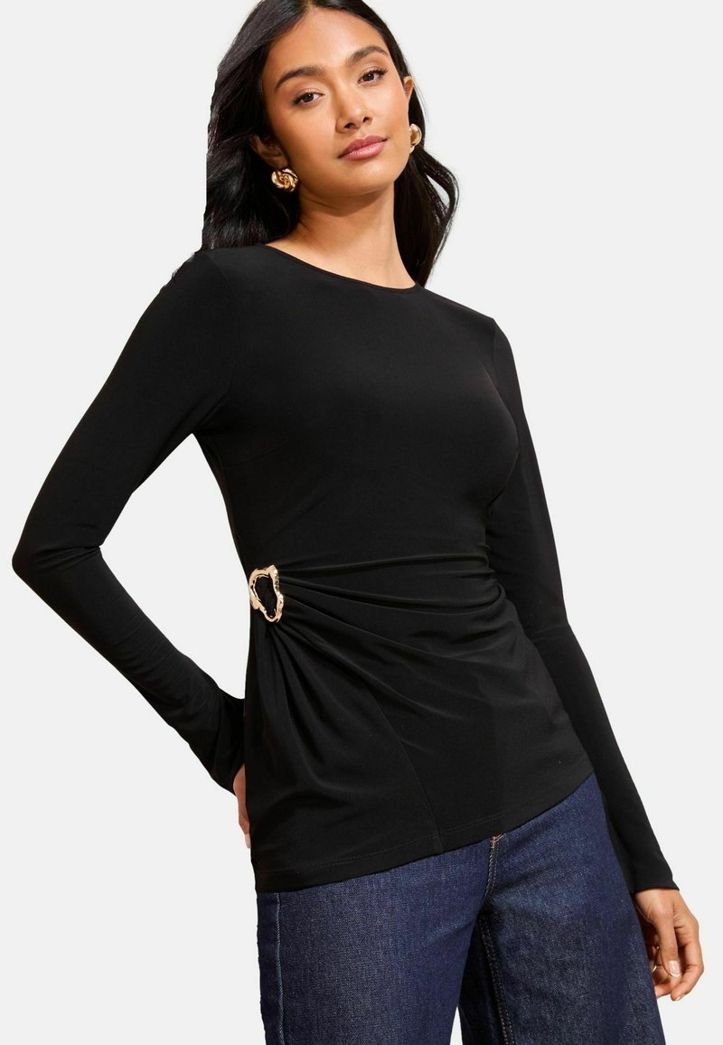 Fekete, hosszú ujjú felső, összegyűrt oldali részletekkel, arany kerek csattal kiemelve. Sima anyag, testhezálló kialakítással. Sötét farmernadrággal párosítva.