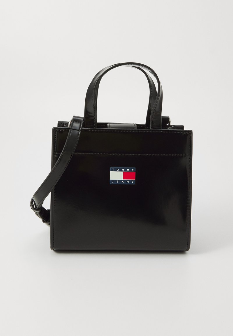 Sac à main en cuir verni noir avec deux poignées supérieures, une bandoulière réglable et un patch logo à l'avant aux couleurs bleu, rouge et blanc.