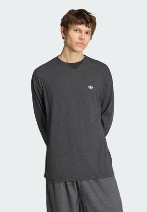 Joven con camiseta de manga larga Adidas gris oscuro y pantalones a juego, de pie con las manos detrás de la espalda contra un fondo liso y claro.