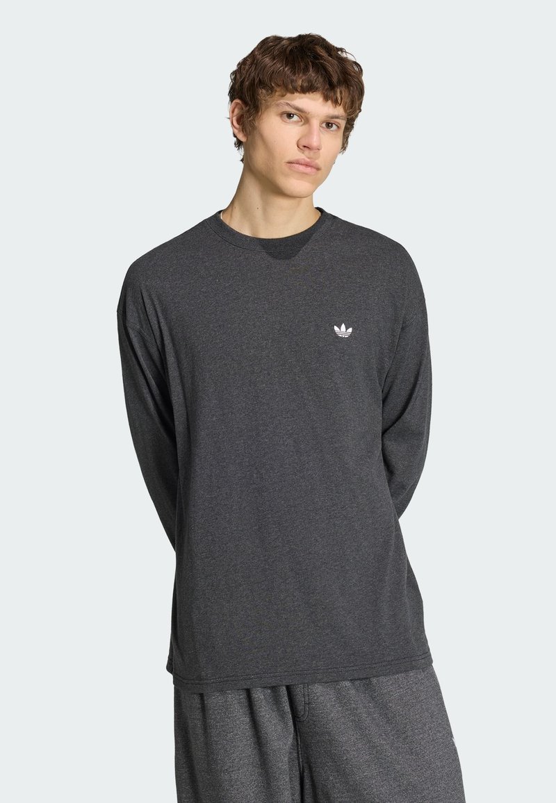 Jeune homme portant un t-shirt Adidas à manches longues gris foncé et un pantalon assorti, debout, les mains derrière le dos, sur un fond clair uni.