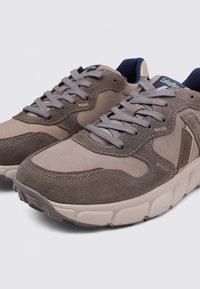 Une paire de baskets en suède gris et beige avec de grosses semelles blanches et des lacets gris, dotées d'un intérieur bleu marine et d'étiquettes de langue de marque.