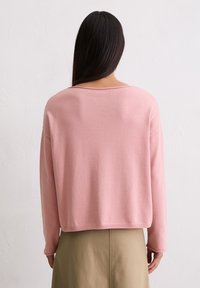 Lichtroze, langemouwtrui met een relaxte pasvorm en ruime halslijn. Zachte textuur, effen kleur, getoond vanuit de achterkant, gecombineerd met een beige rok.