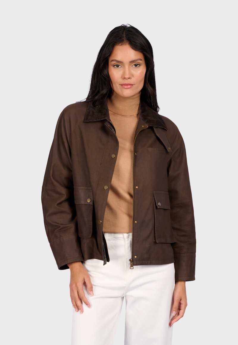 Hnědá kožená bunda s měkkým semišovým límcem, s knoflíkovými kapsami a volným střihem. Nositá přes béžový rolák.